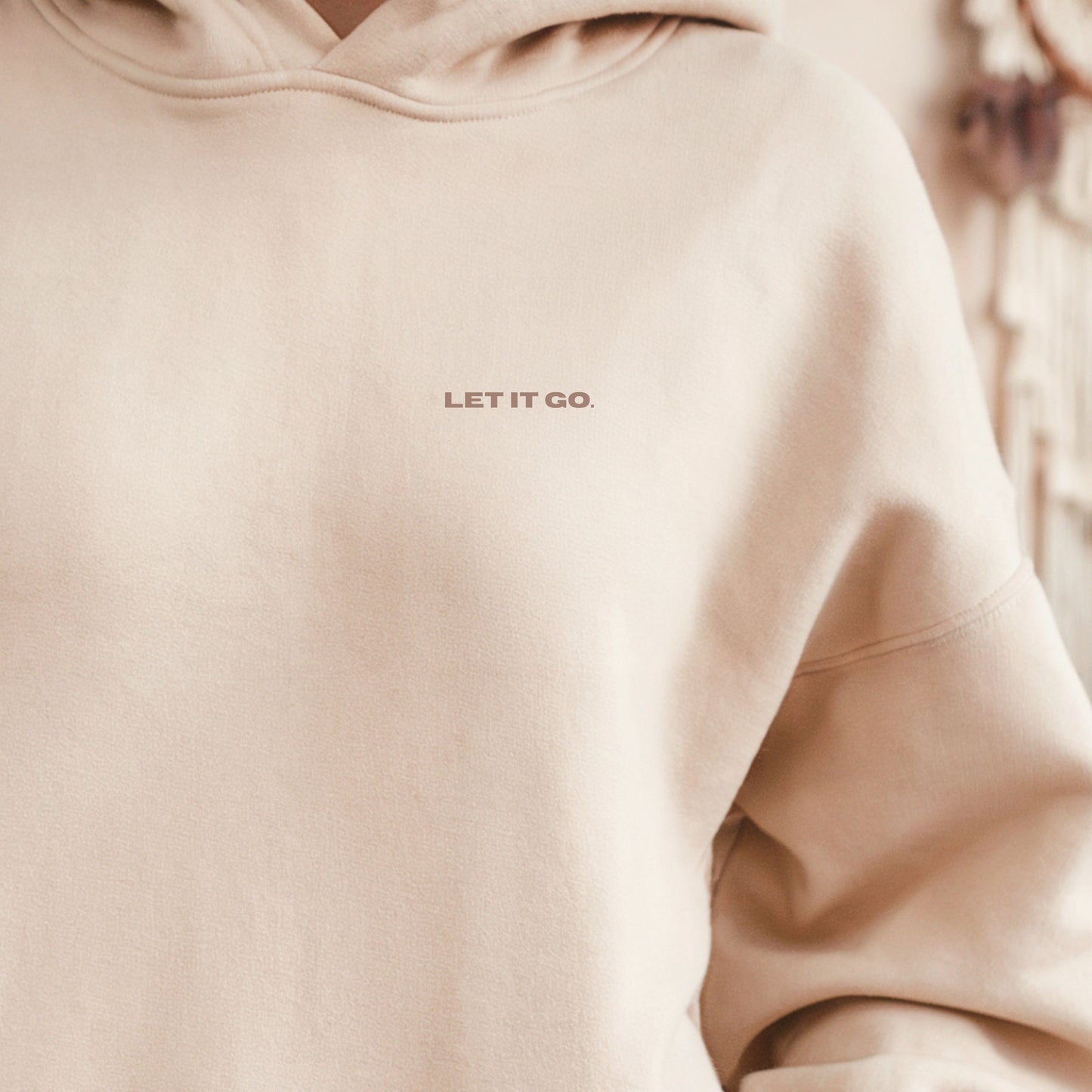 Model wearing the sand VIKA & EVE “LET IT GO” premium organic hoodie indoors wit a cofee— minimal unisex design with a soft, cozy heavyweight feel.