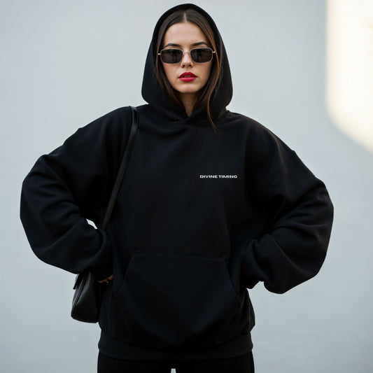 Photo with model wearing black oversized VIKA & EVE DIVINE TIMING hoodie by the water – minimalist unisex design for who we strive to be.
