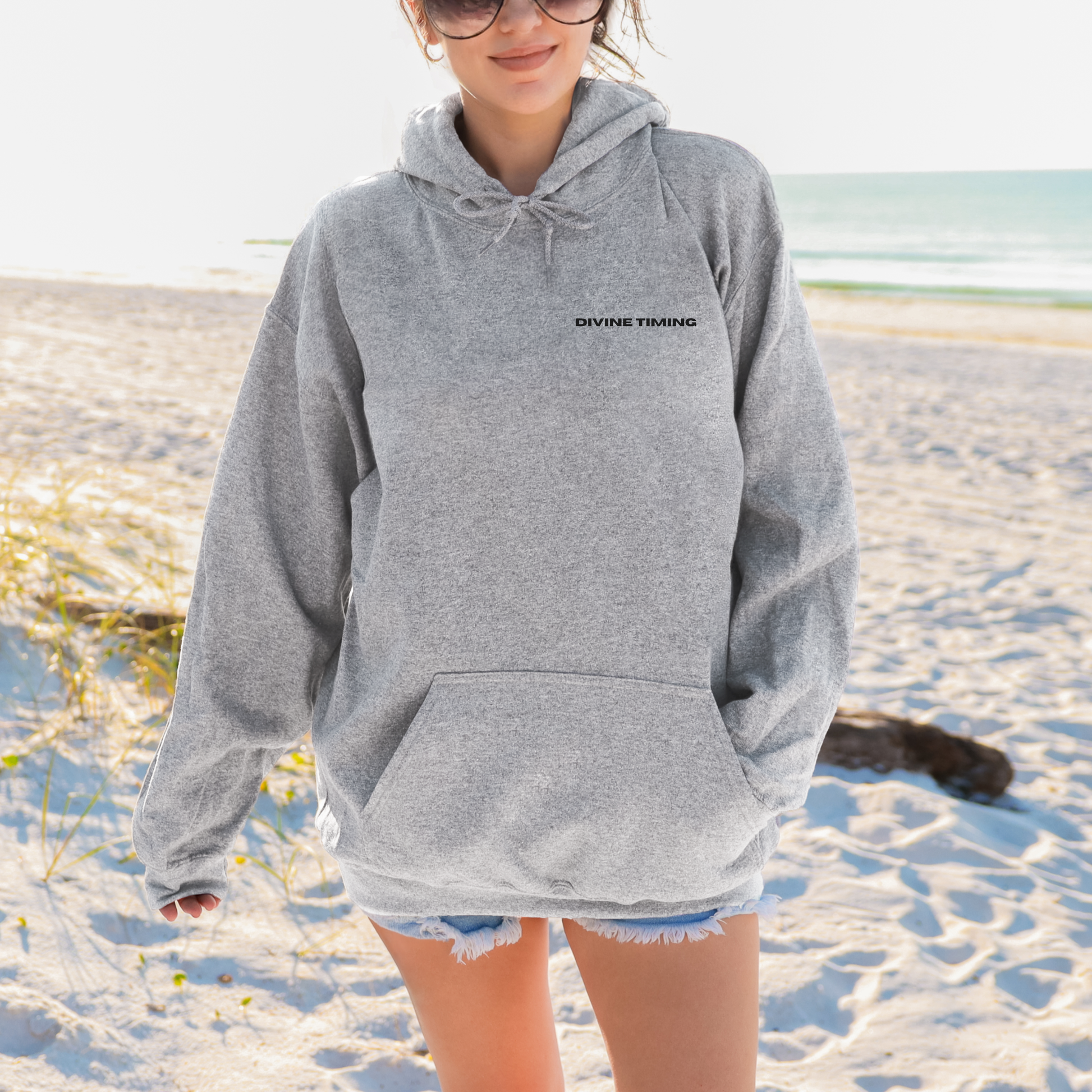 Model wearing grey oversized VIKA & EVE  DIVINE TIMING hoodie on a beach – minimalist unisex design grounded in trust and divine flow.
