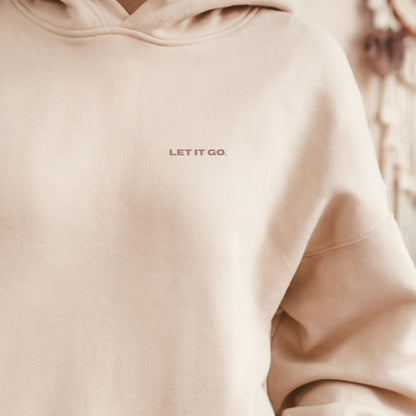 Model wearing the sand VIKA & EVE  “LET IT GO” premium organic hoodie indoors wit a cofee— minimal unisex design with a soft, cozy heavyweight feel.