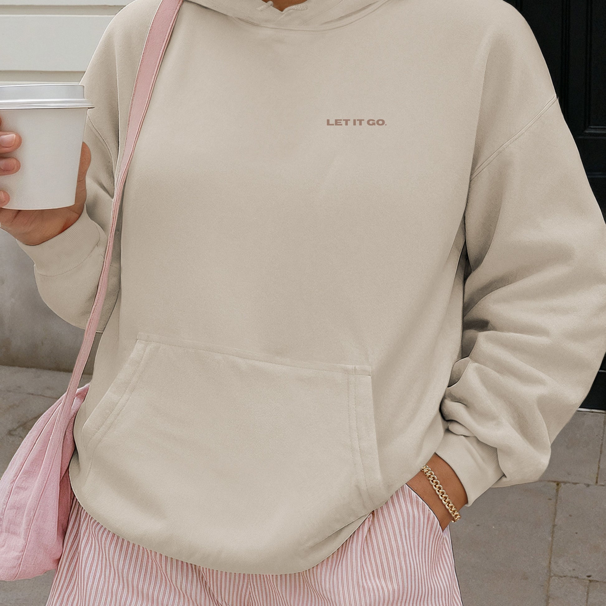 Model wearing the sand hoodie VIKA & EVE  “LET IT GO” premium organic hoodie indoors wit a cofee— minimal unisex design with a soft, cozy heavyweight feel.