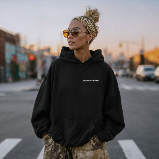Model wearing black oversized VIKA&EVE  DIVINE TIMING hoodie at sunset in the city – minimalist unisex design symbolizing trust, timing, and calm presence.