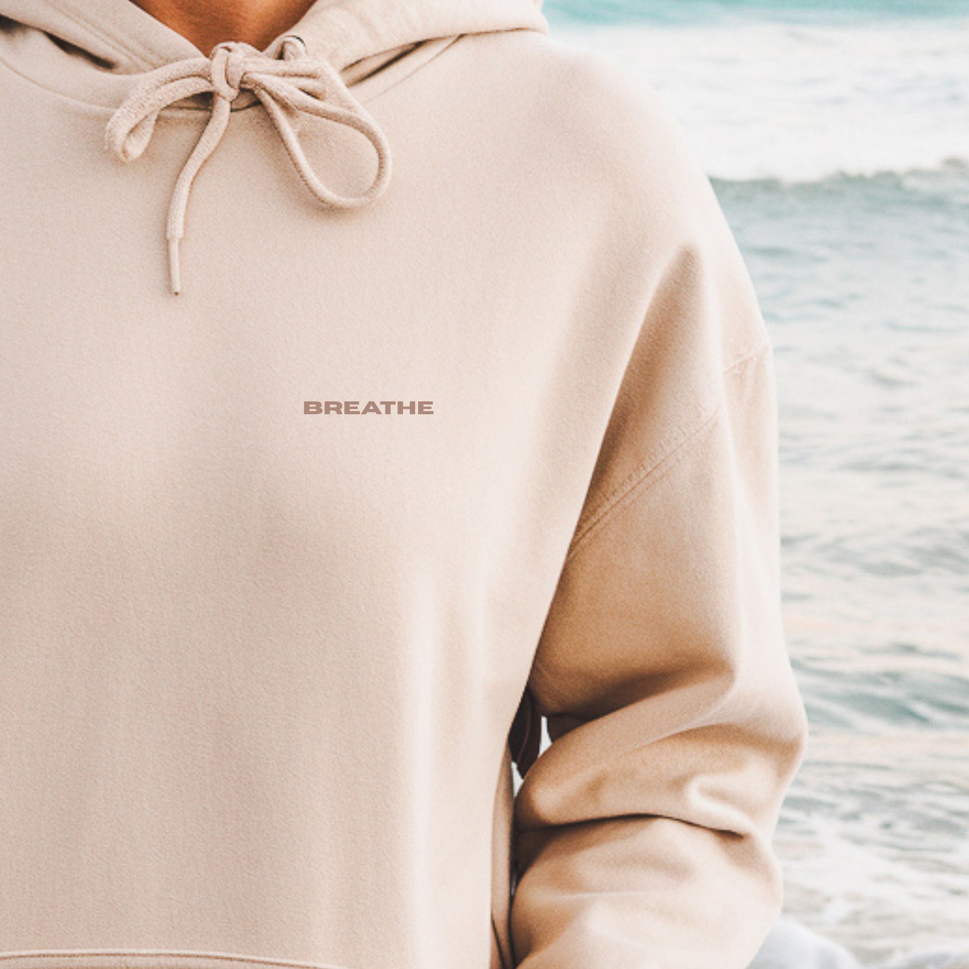 Close-up of model wearing sand oversized Vika&Eve  BREATHE hoodie by the water – minimalist unisex design for who we strive to be.